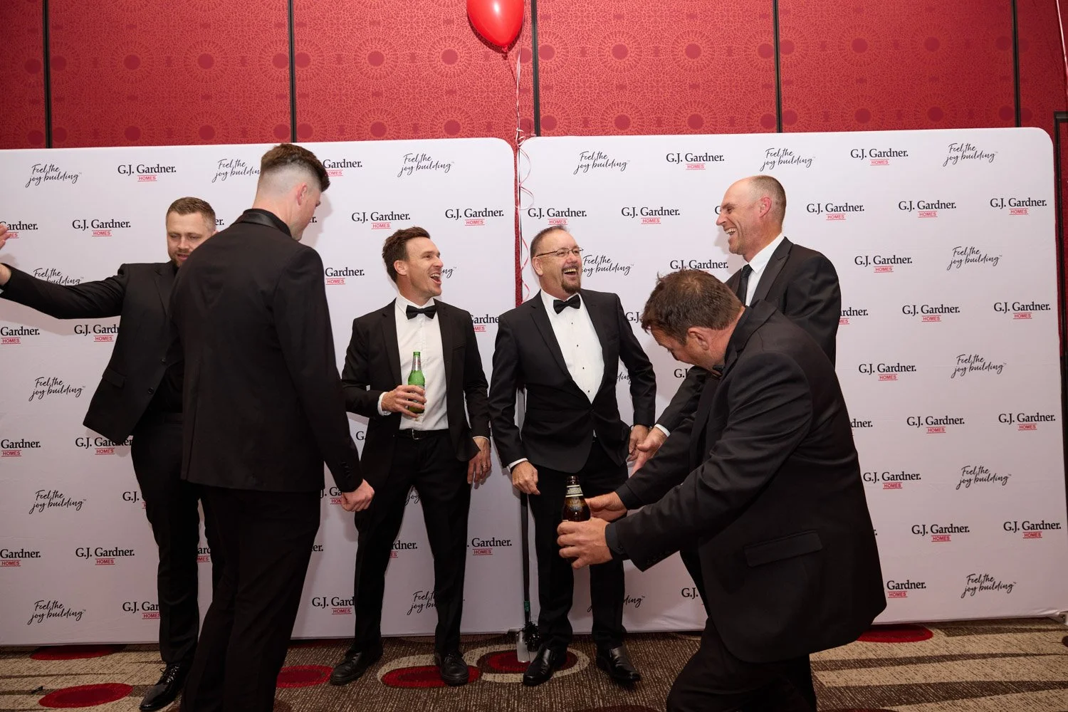 Group of men in tuxedos and suits, laughing and holding drinks, at an event with a G.J. Gardner Homes backdrop.