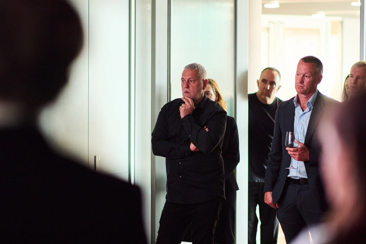 Group of professionally dressed people at a reception or event, standing indoors near glass doors, engaging in conversation or listening attentively.