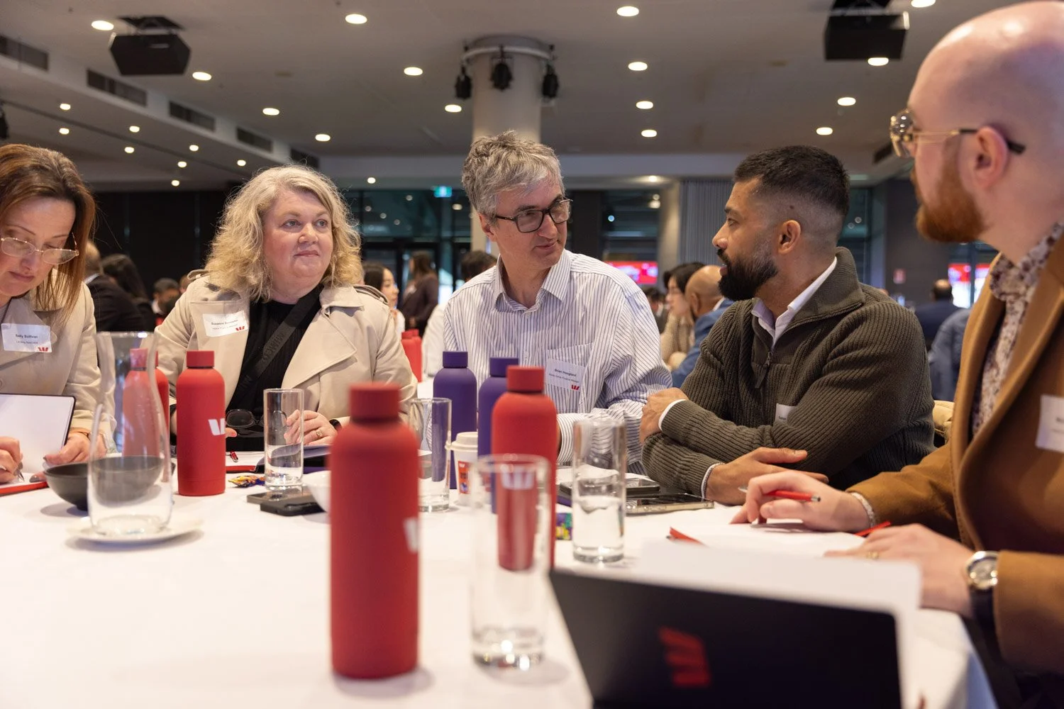 Group of five people sitting around a table engaged in conversation at a conference or business event indoors.