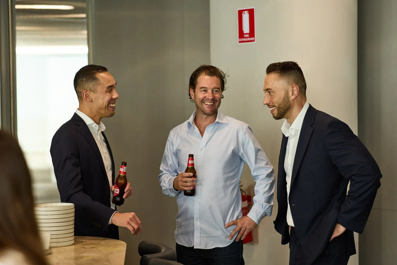 Three men in business attire holding beer bottles engaging in conversation and smiling at each other in an indoor setting.