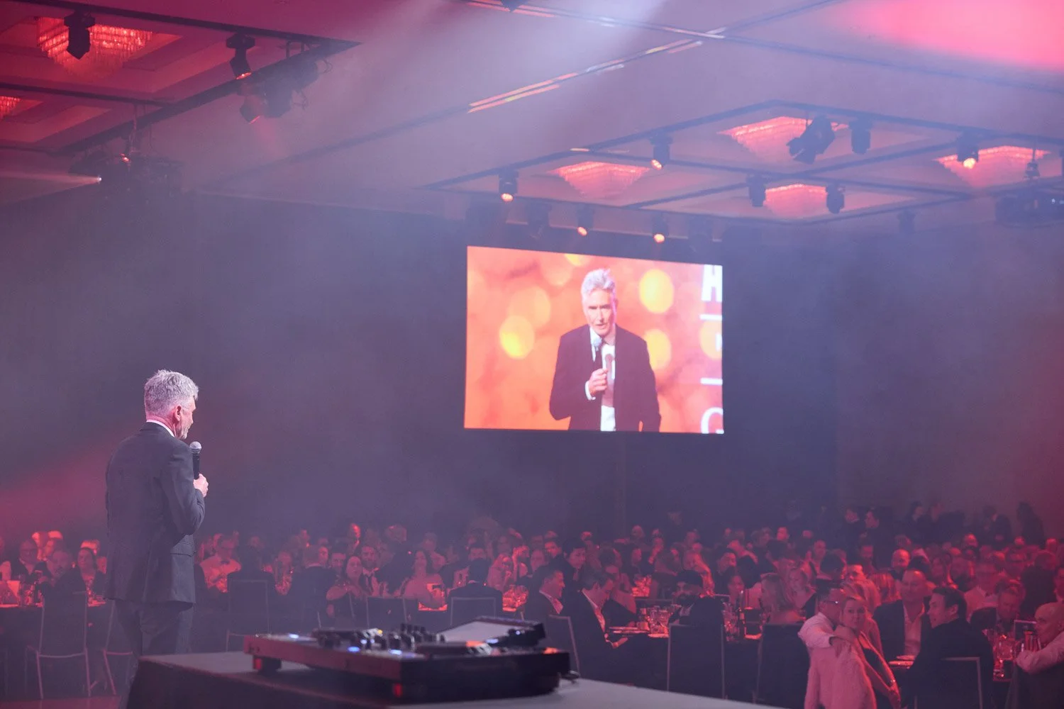 Man in a suit speaking into a microphone at a large formal event, with many attendees seated at tables in the background, and his image projected on a large screen behind him.