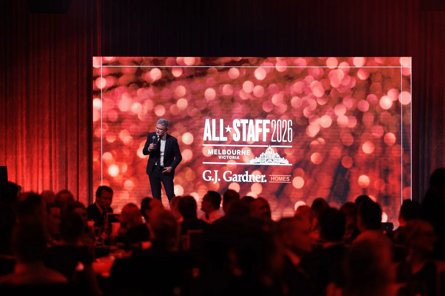 A man in a dark suit giving a speech on stage at an event with a large screen behind him displaying the event name 'ALL STAFF 2026' and logos for Melbourne, Victoria, G.J. Gardner Homes. The audience is seated in front, with red curtains framing the 