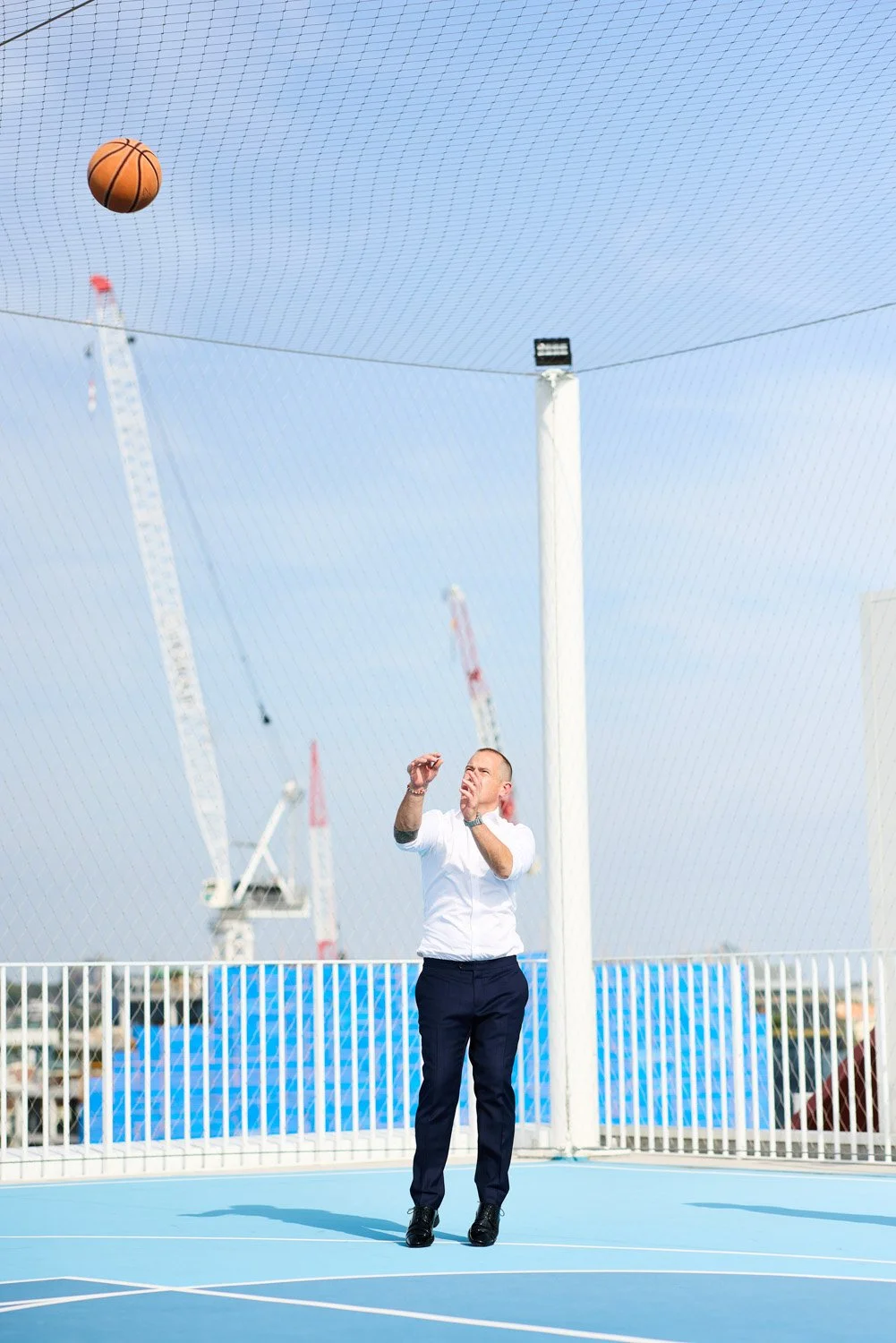 A man in business attire standing on a blue outdoor basketball court, shooting a basketball towards the hoop under a clear sky.