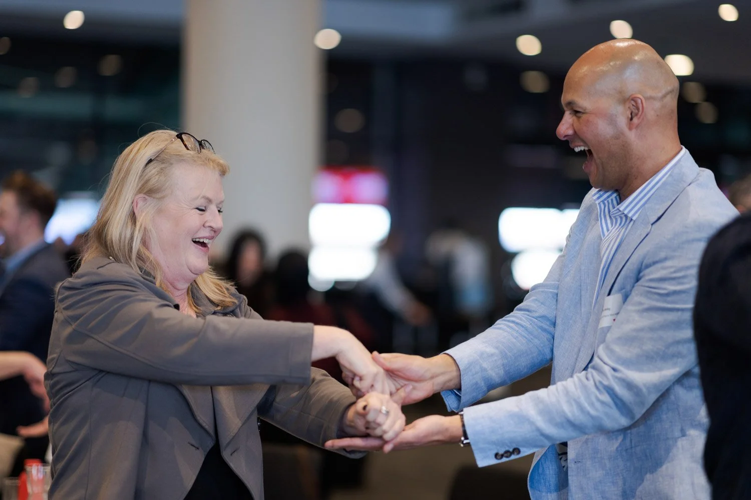 Two people, a woman and a man, are smiling and shaking hands at an indoor event. The woman has blonde hair, glasses on her head, and is wearing a brown blazer. The man is bald, wearing a light blue blazer and a striped shirt. Both are happy and engag