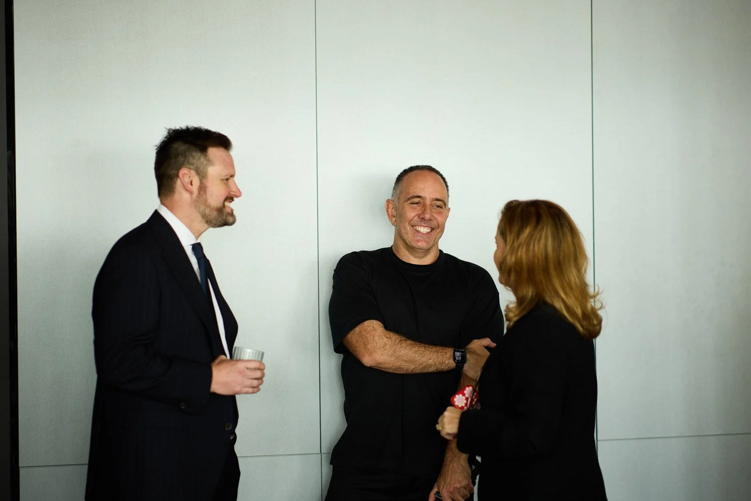 Three people in a professional setting, two men and one woman, engaged in conversation, with one person holding a glass and smiling.