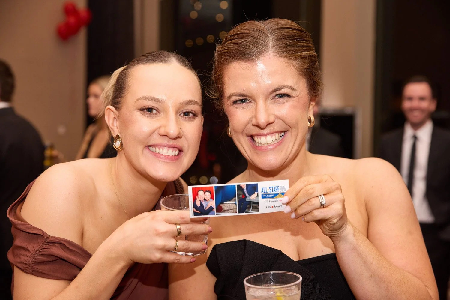 Two women smiling at a celebration event, holding a ticket with photo and event details, with others in the background.