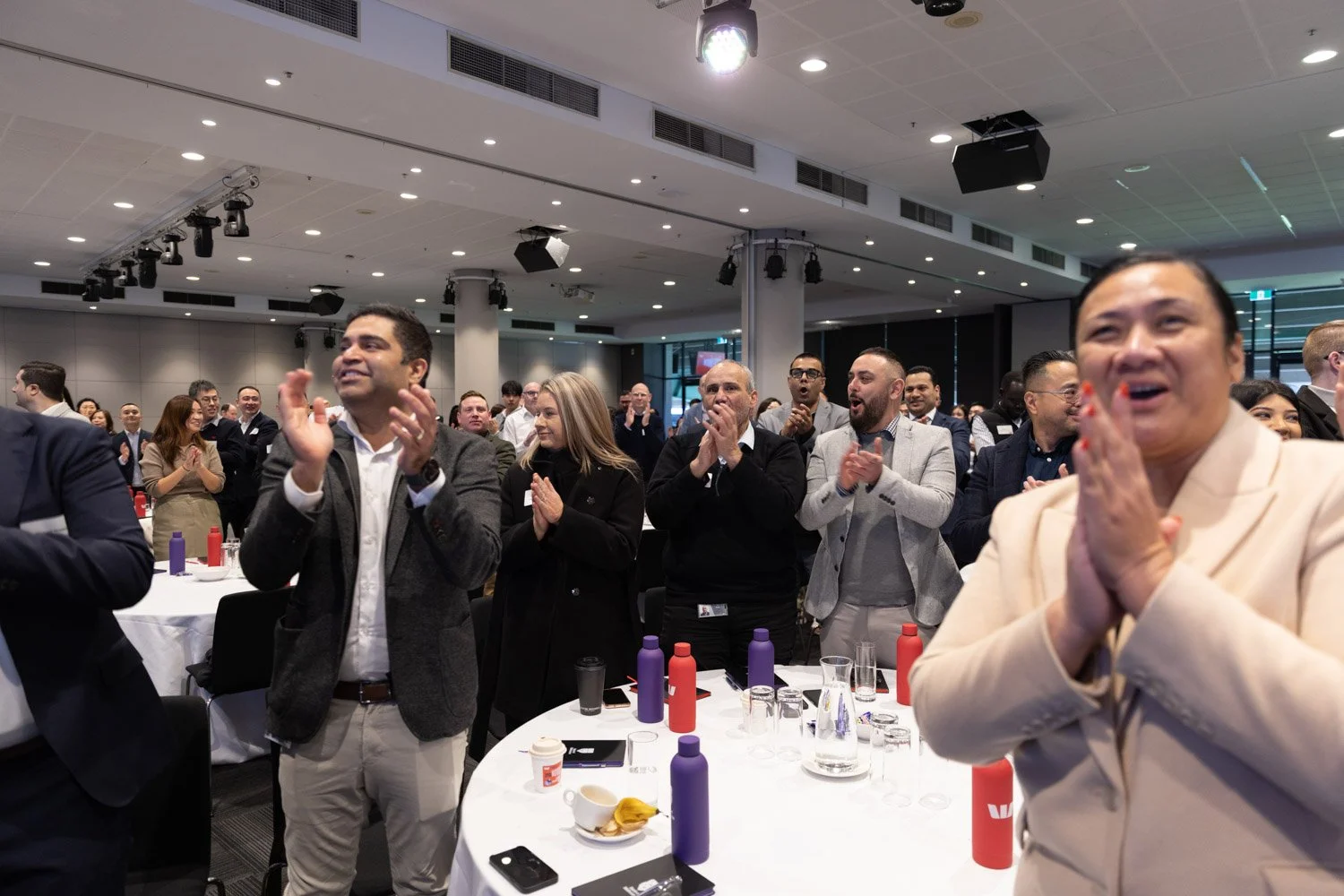 A diverse group of professionals standing and clapping at a conference, with some smiling and attentively watching a speaker or presentation in a modern conference room.