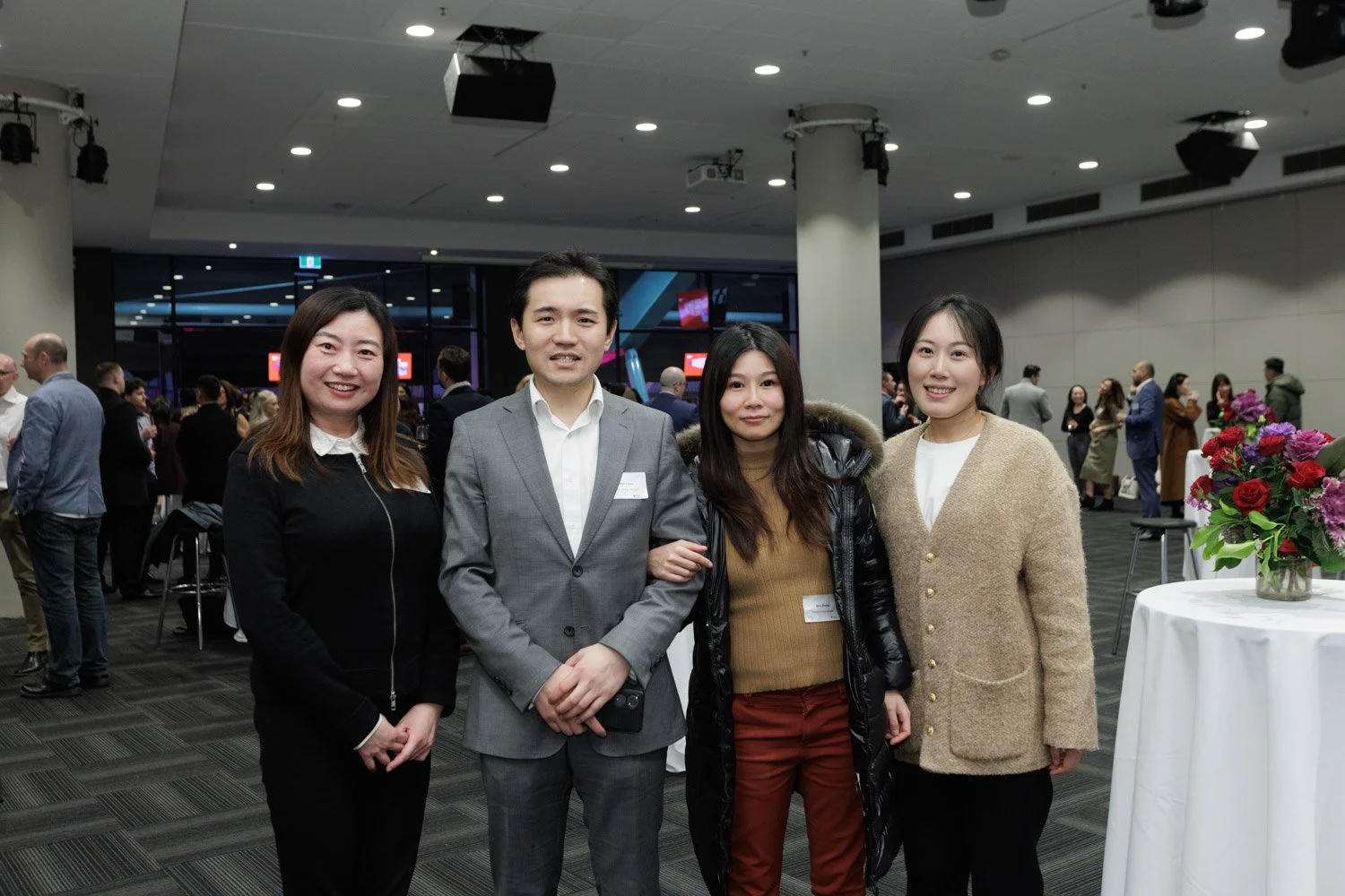 Four people standing together at a conference or event, smiling at the camera, with a group of people in the background and a flower arrangement on a table to the right.