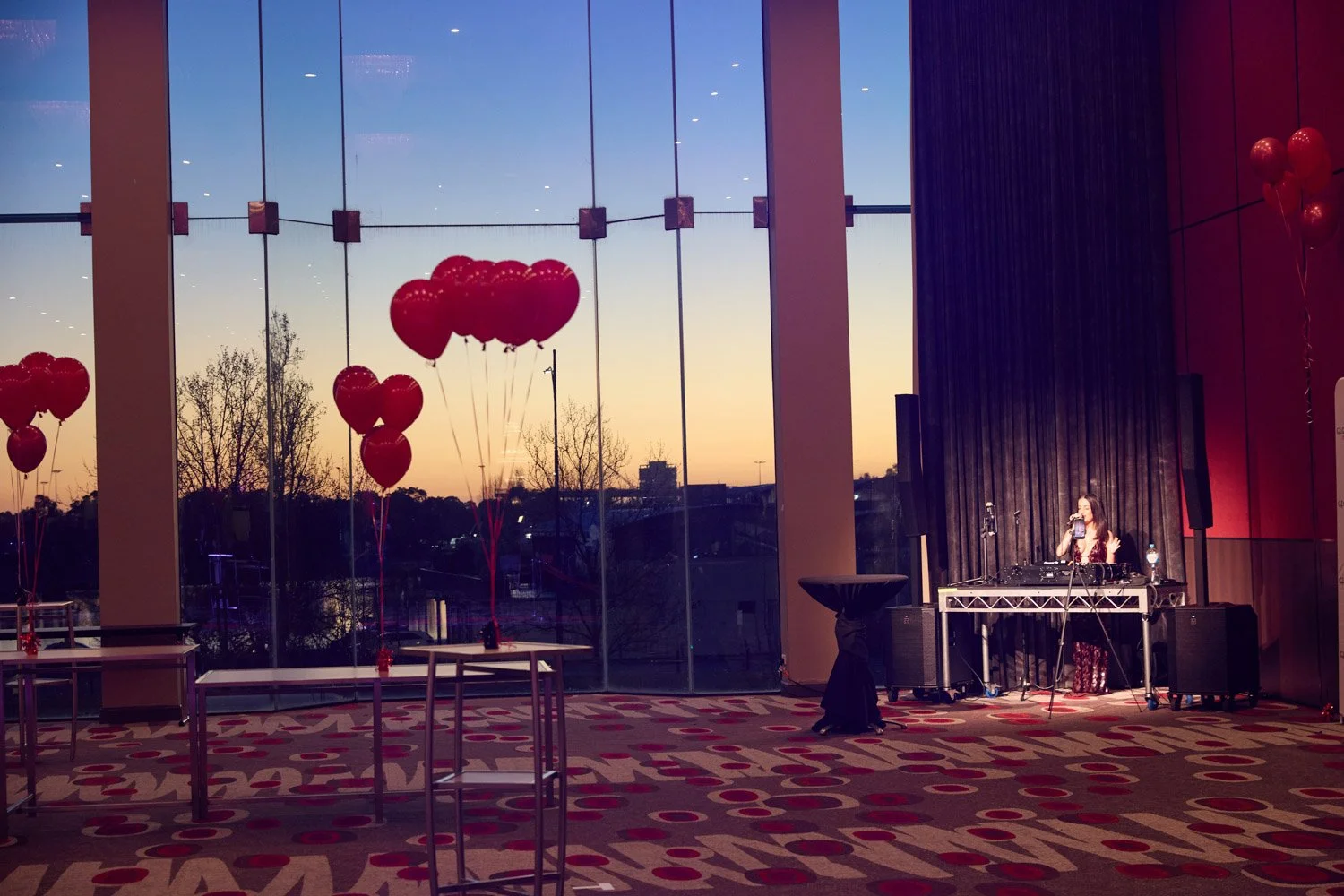 An indoor event space with large windows view of sunset sky and trees outside. Inside, there are red heart-shaped balloons, a DJ or singer performing at a table with sound equipment, and a patterned carpet.