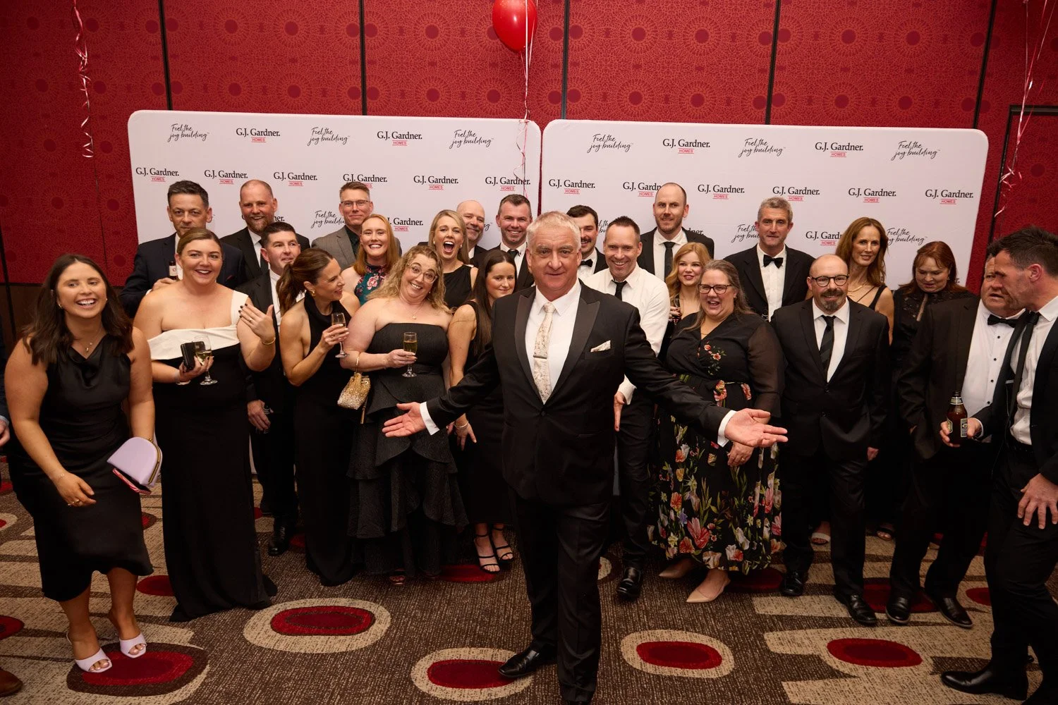 Group of people dressed in formal attire at a celebration event, posing in front of a backdrop with G.J. Gardner Homes branding.