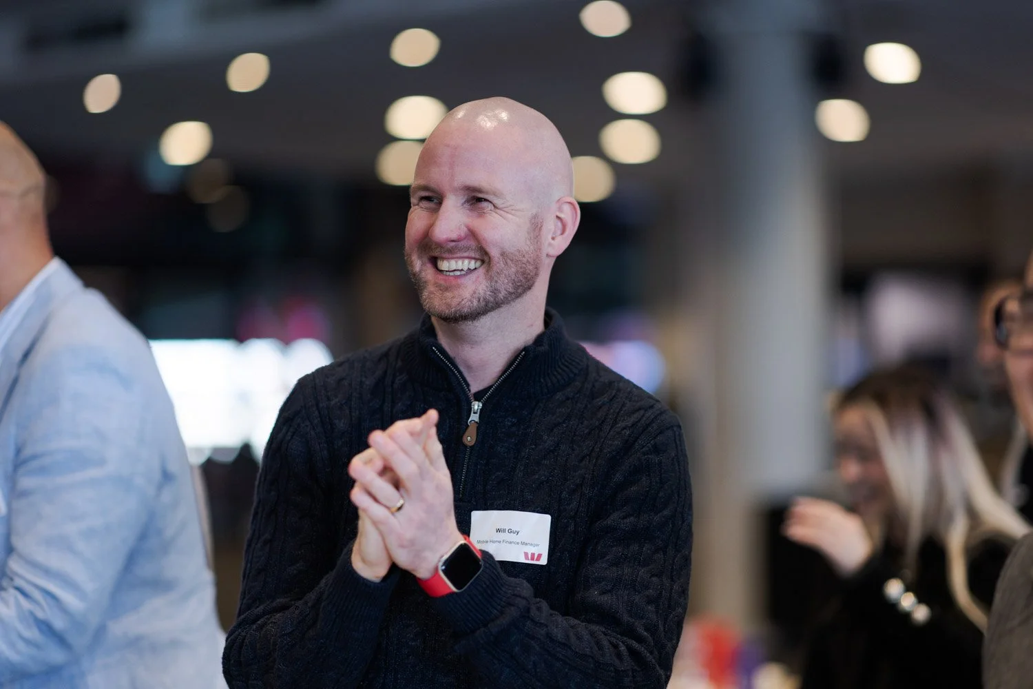 Man smiling, wearing a black cable knit sweater and a smartwatch, at a social event.
