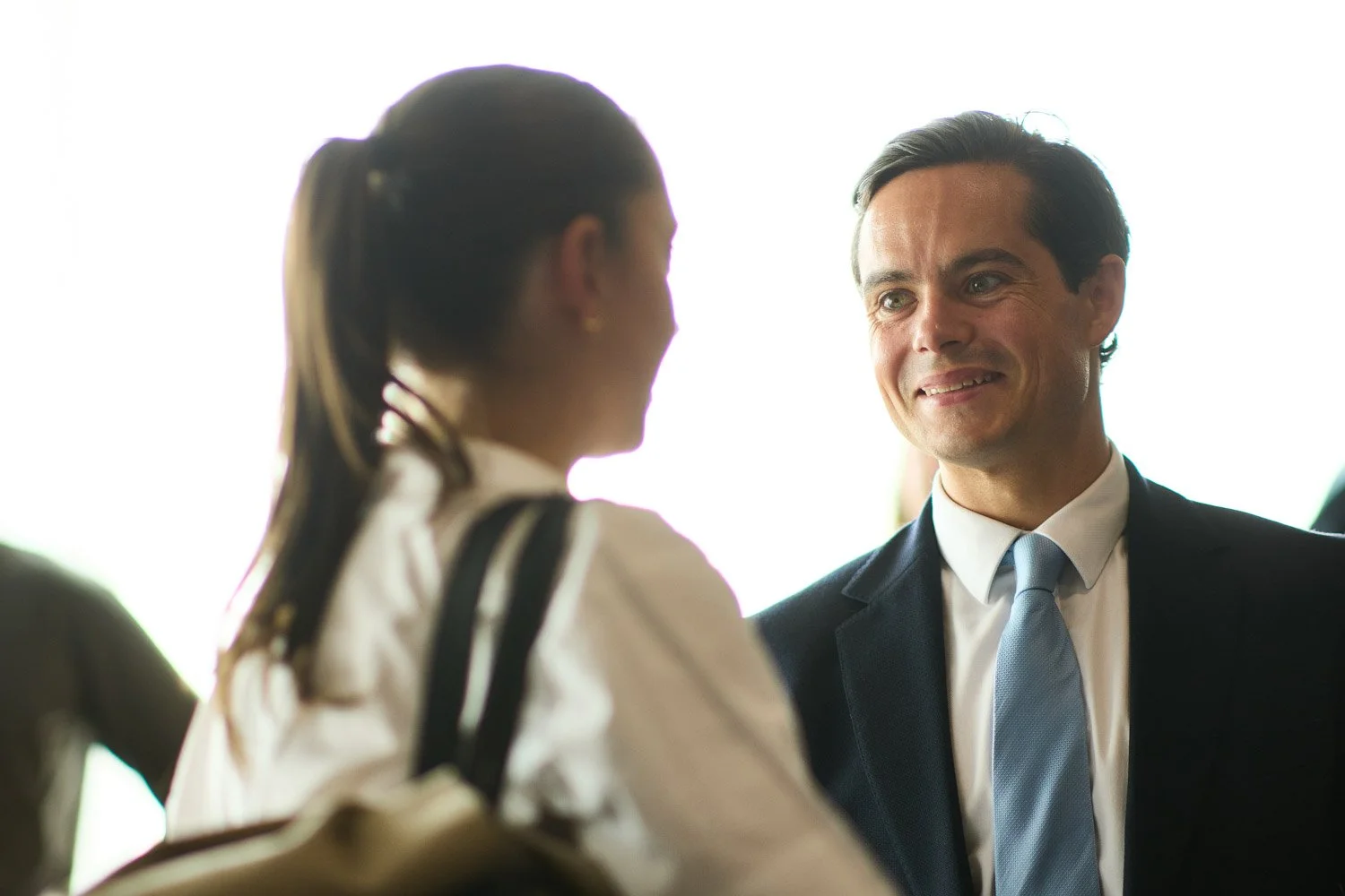 A man in a suit smiling and talking to a woman with her back to the camera, in a bright indoor setting.