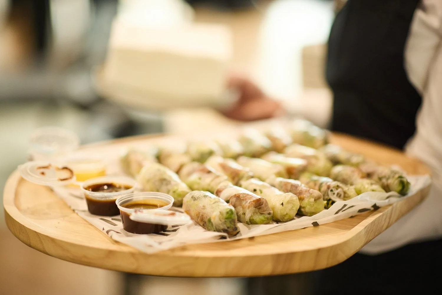 A server holds a wooden tray with fresh vegetable spring rolls and small cups of dipping sauce