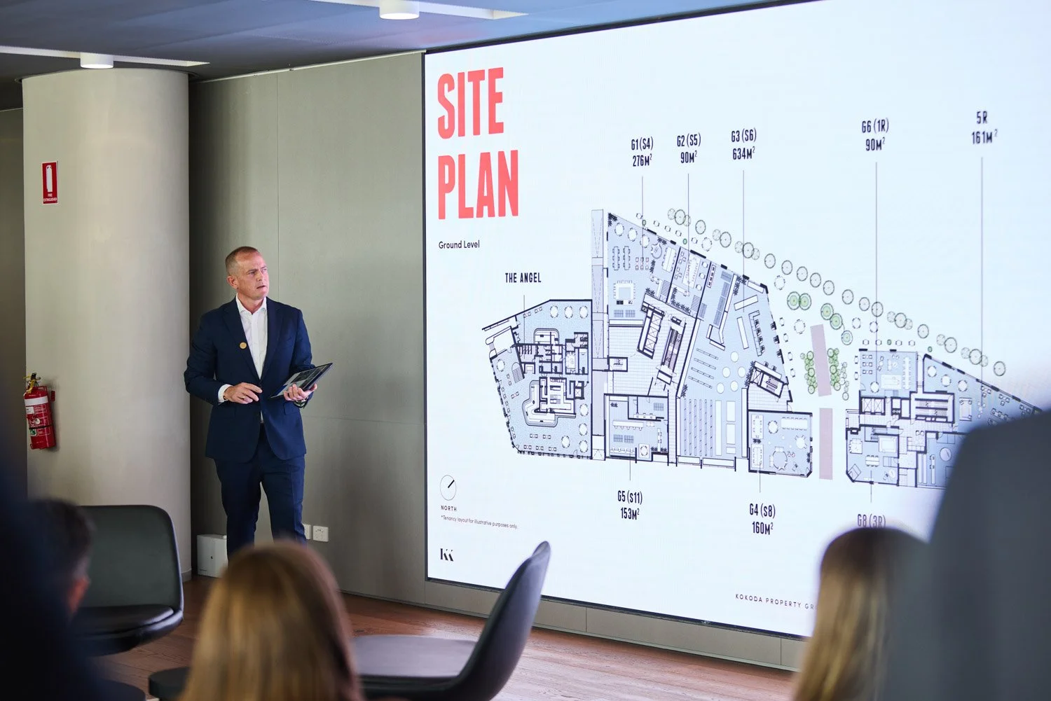 A man in a navy suit giving a presentation in a conference room, standing next to a large screen displaying a site plan with labeled sections and floor areas.