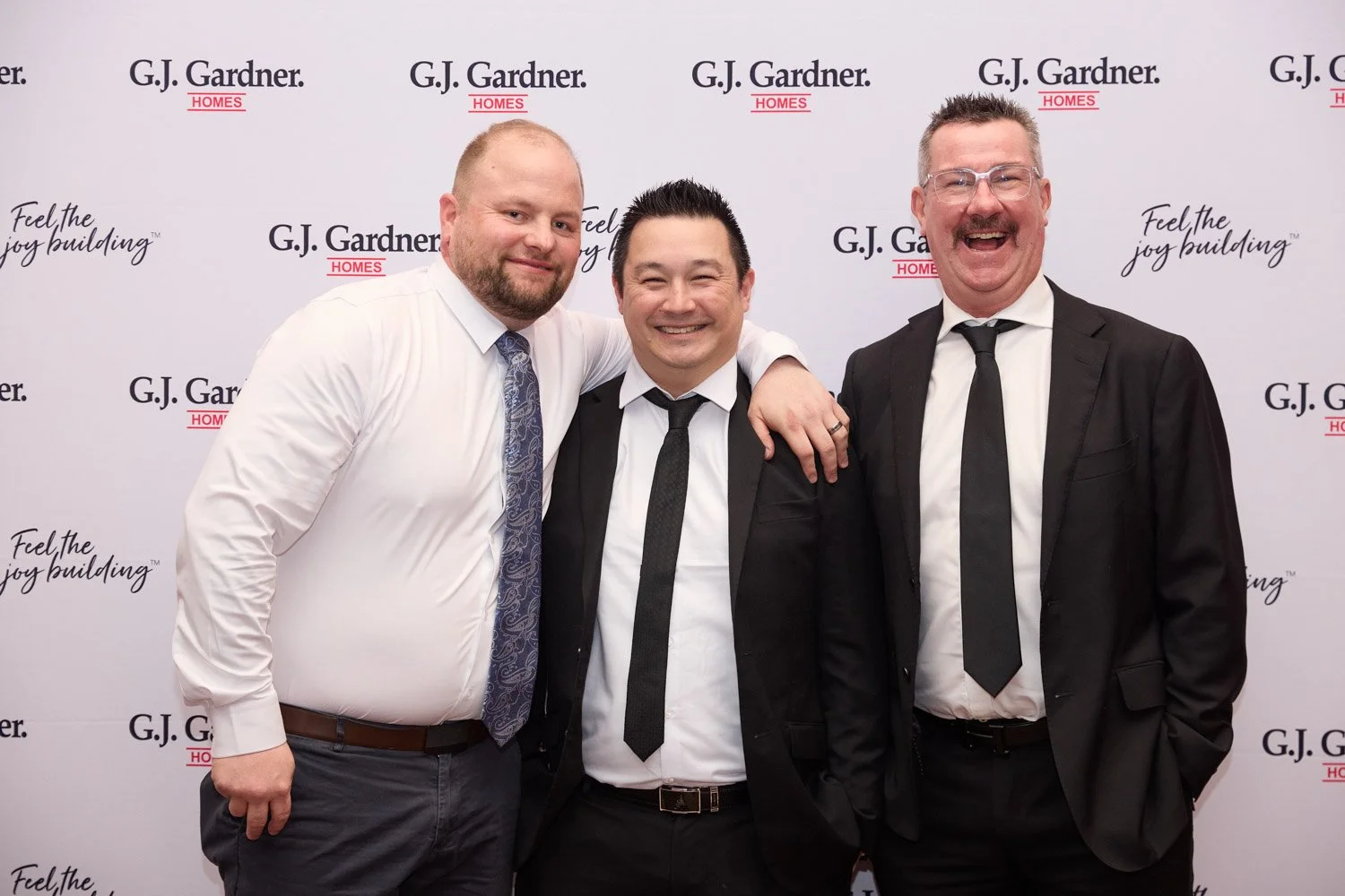 Three men in formal attire posing together at an event, with a backdrop featuring the text "G.J. Gardner Homes" and "Feel the joy building."
