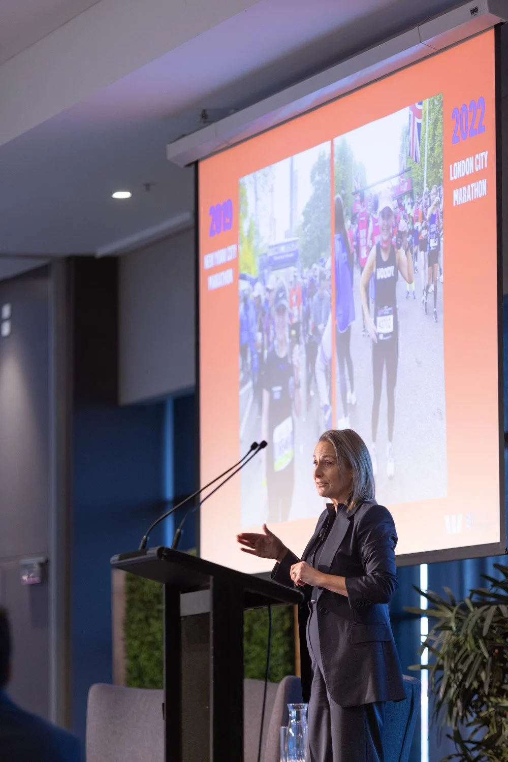 A woman speaking at a podium during a presentation, with a large screen displaying images of a marathon race in London City in 2022 behind her.