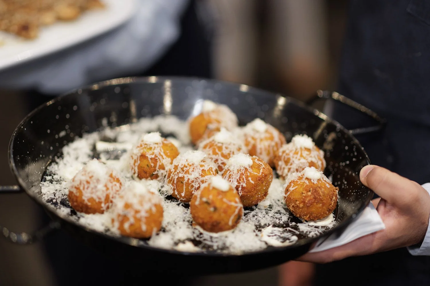 A hand holding a black tray with fried food balls topped with shredded cheese and sauce, with a plate of similar food in the background.
