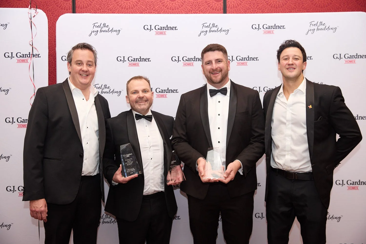 Four men in tuxedos standing together at an award ceremony, holding glass awards.