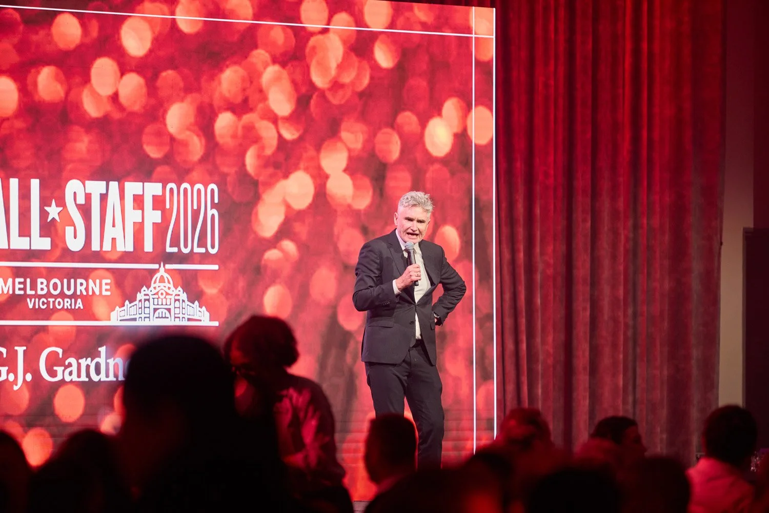 A man in a black suit standing on stage, speaking into a microphone, during a formal event in Melbourne, Victoria, 2020. The stage background features red and orange bokeh lights with event text and a building illustration.