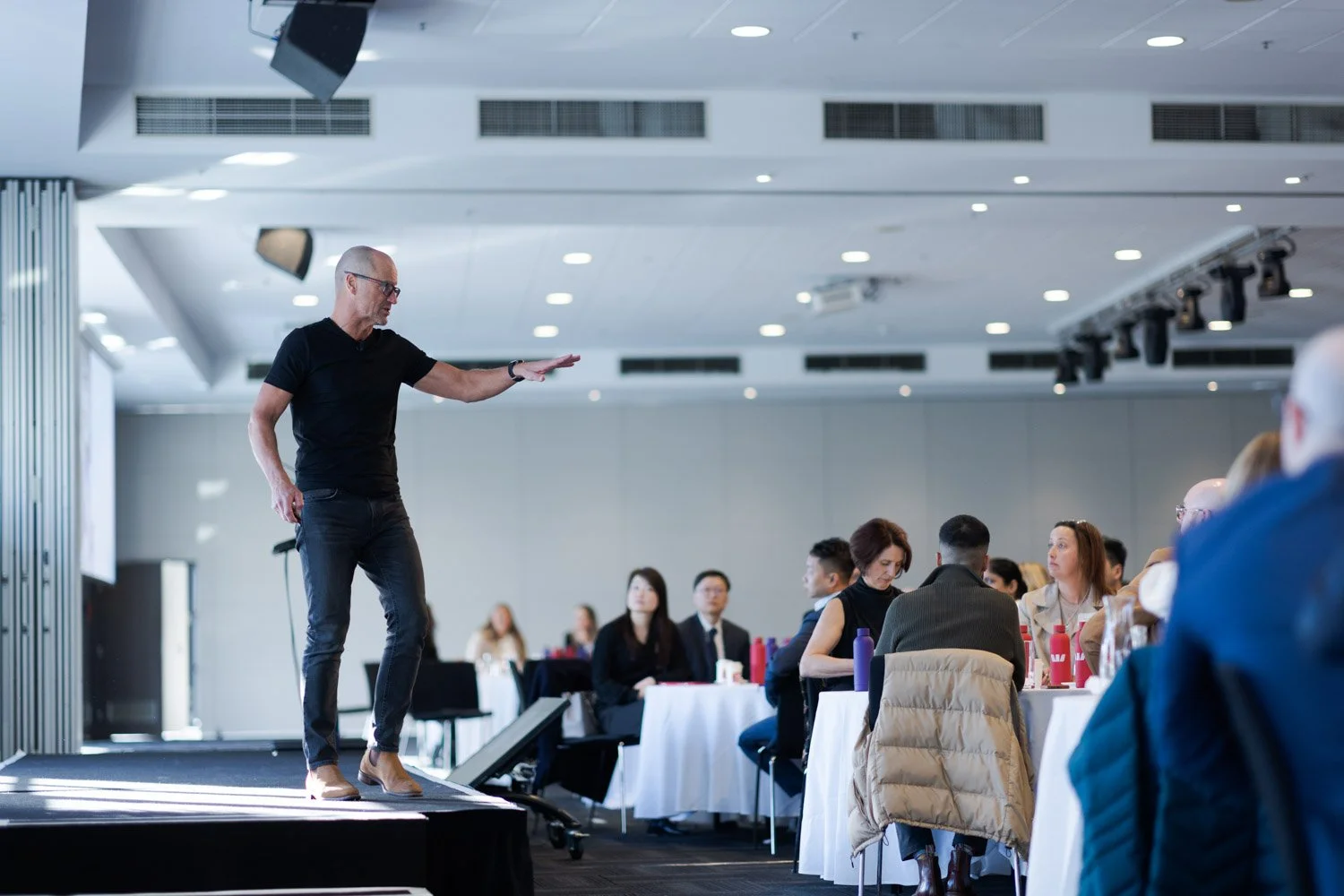 A man giving a presentation or speech on a stage in front of an audience seated at tables during a conference or seminar in a modern, well-lit room.