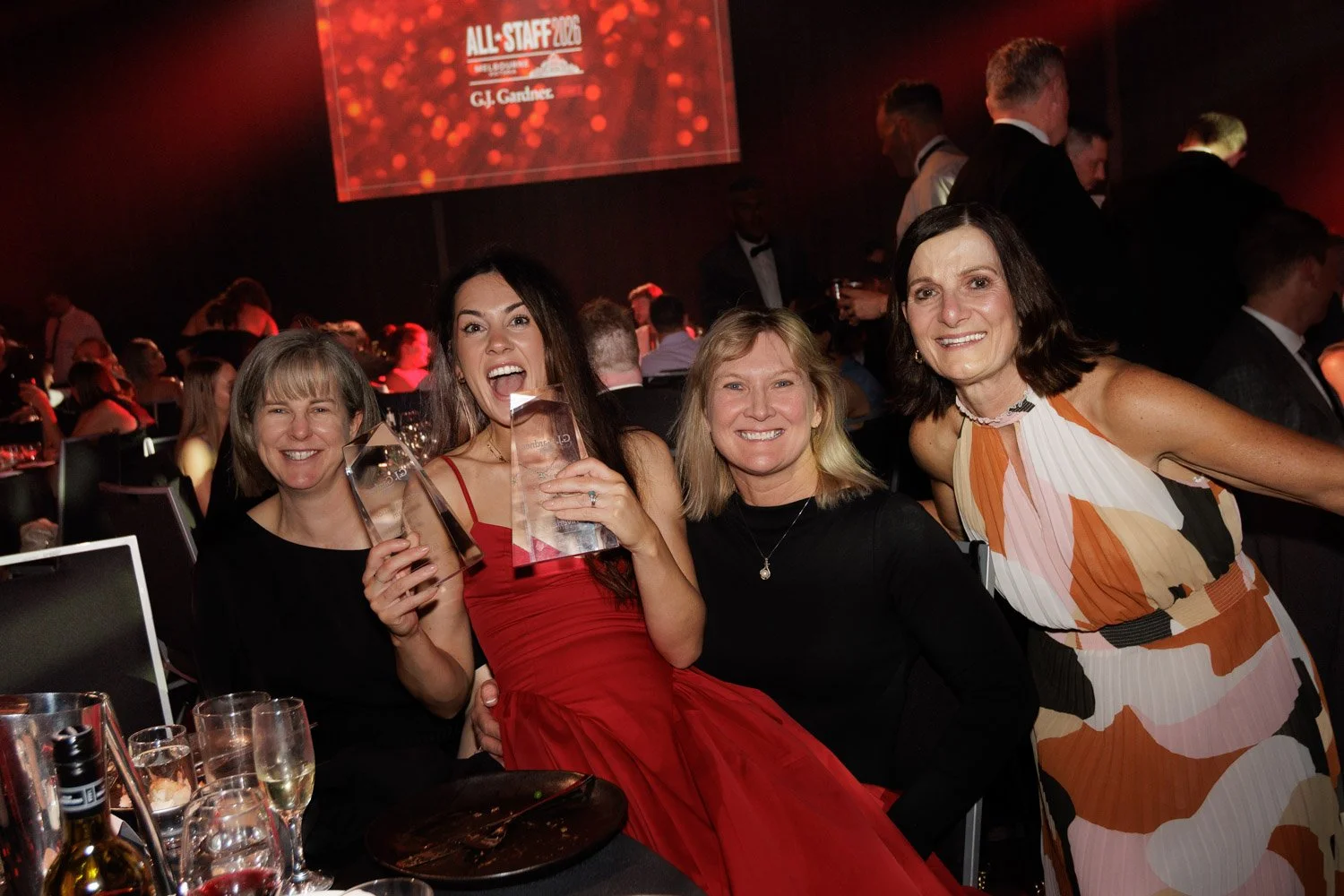 Four women at an awards ceremony celebrating with drinks, some holding trophies, smiling, with a red-themed background and a large screen displaying awards information.