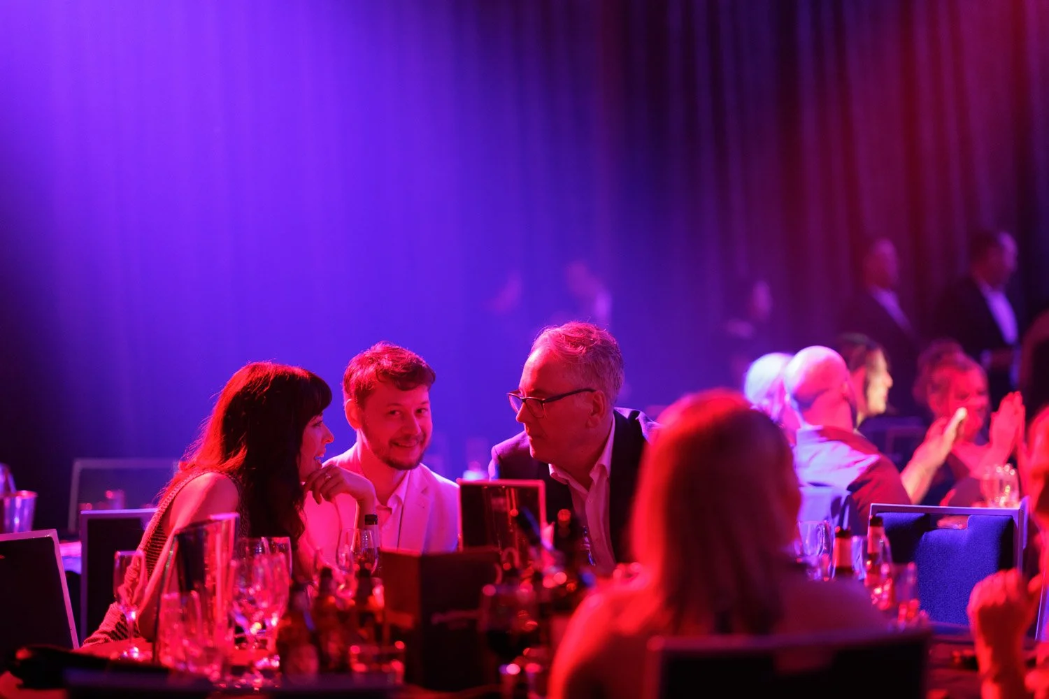 People socializing at a formal event with purple and pink lighting, seated at tables with glassware and bottles.