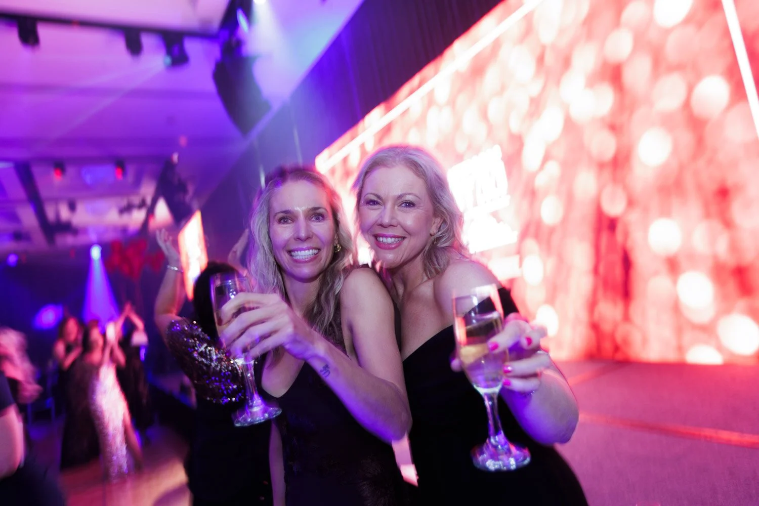 Two women in black dresses smiling and holding glasses of champagne at a party with colorful lights and a red patterned background.