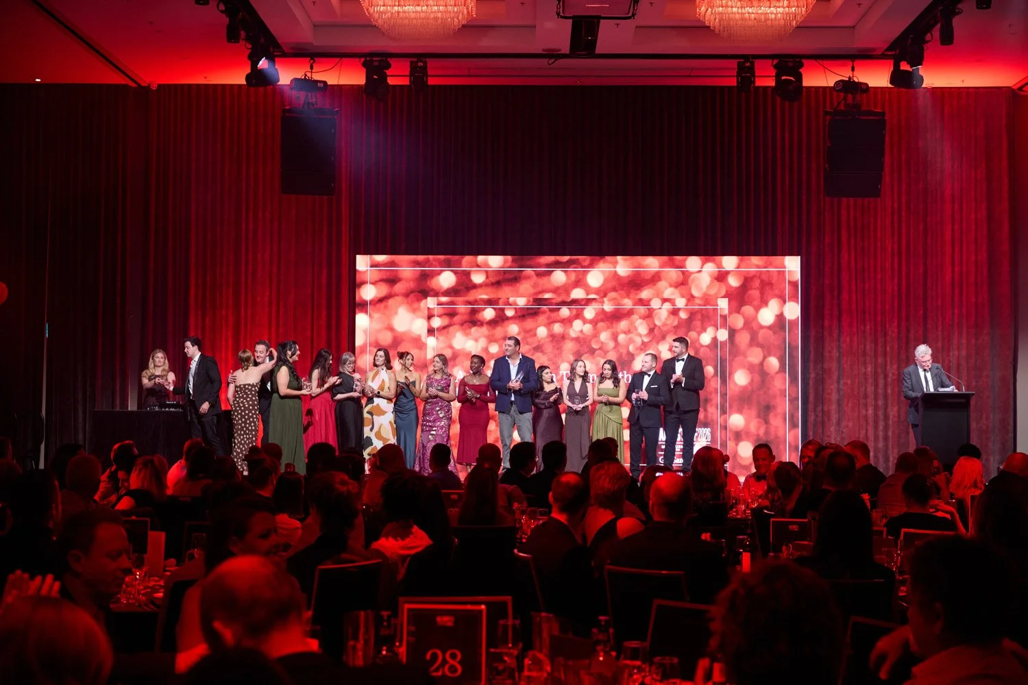 People on stage at an awards ceremony, with a presenter at a podium on the right, large digital screen showing blurred lights, and an audience seated at tables in a red-themed ballroom.