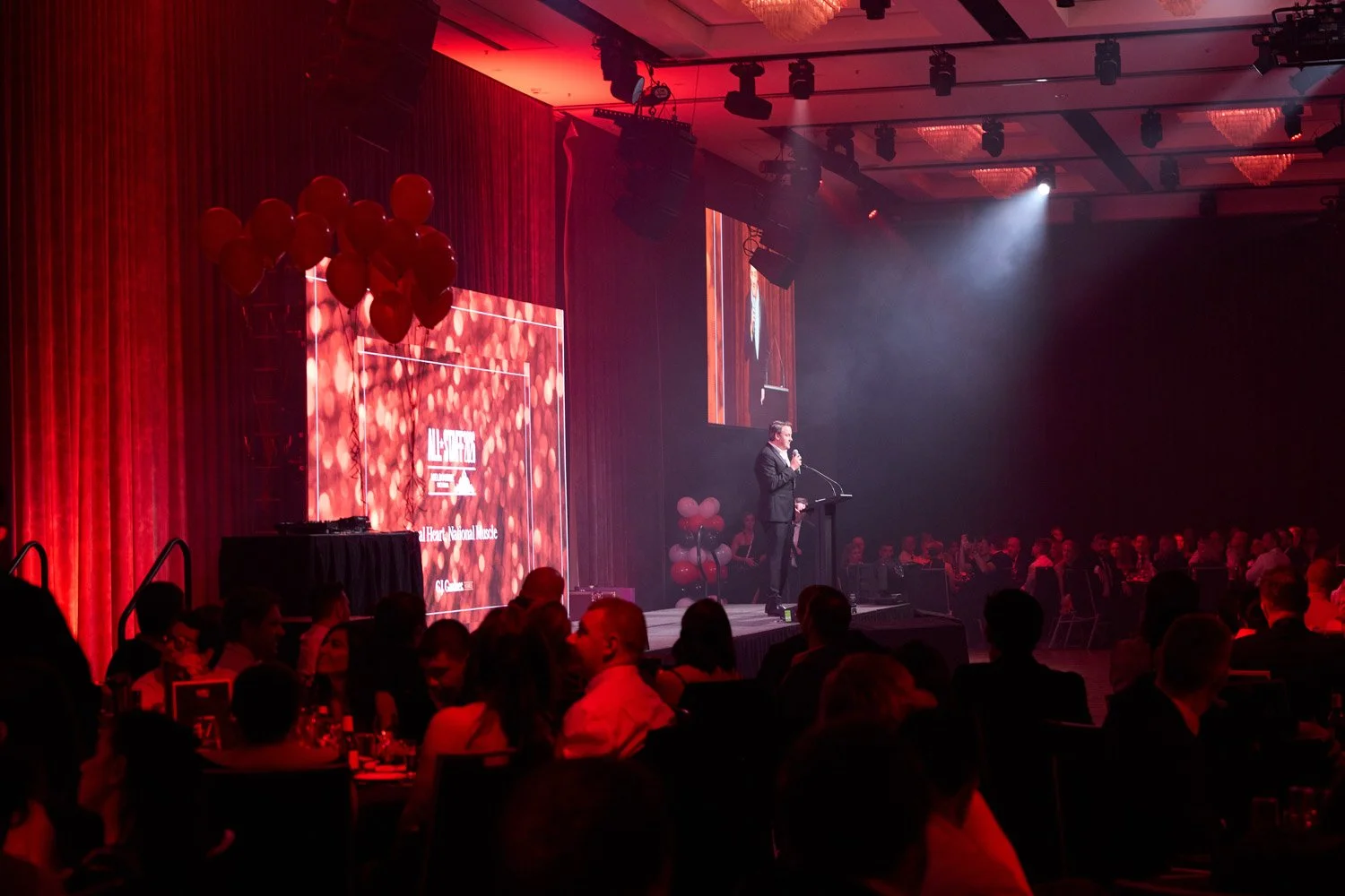 A man in a suit speaking at a podium on a stage with a large screen displaying 'ALL-STATE' and 'South All-State' during a formal event. The stage is decorated with red balloons and red curtains, while the audience seated at tables watches the present