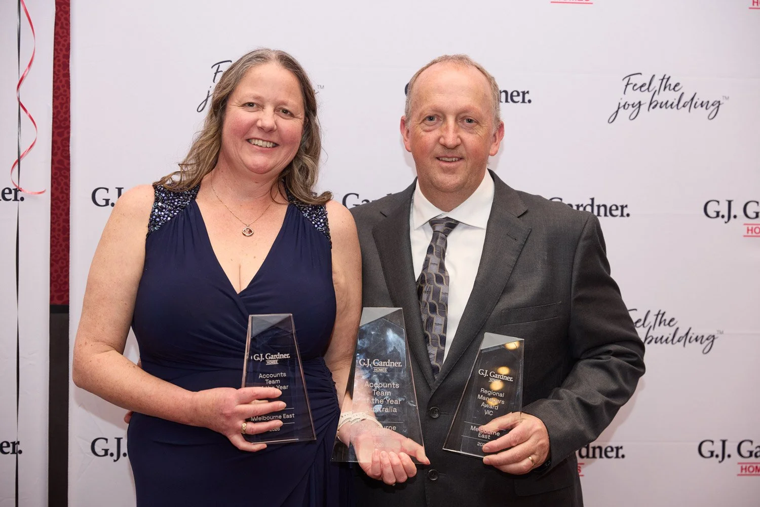 Man and woman in formal attire holding awards, standing in front of a step-and-repeat banner with G.J. Gardner Homes logo and slogan. Woman in blue dress, man in black suit with patterned tie, smiling.