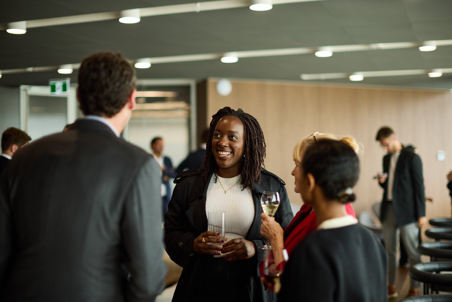 Four people are engaged in conversation at a professional or social event, holding drinks in a modern, well-lit room with a wood-paneled wall and ceiling lights.
