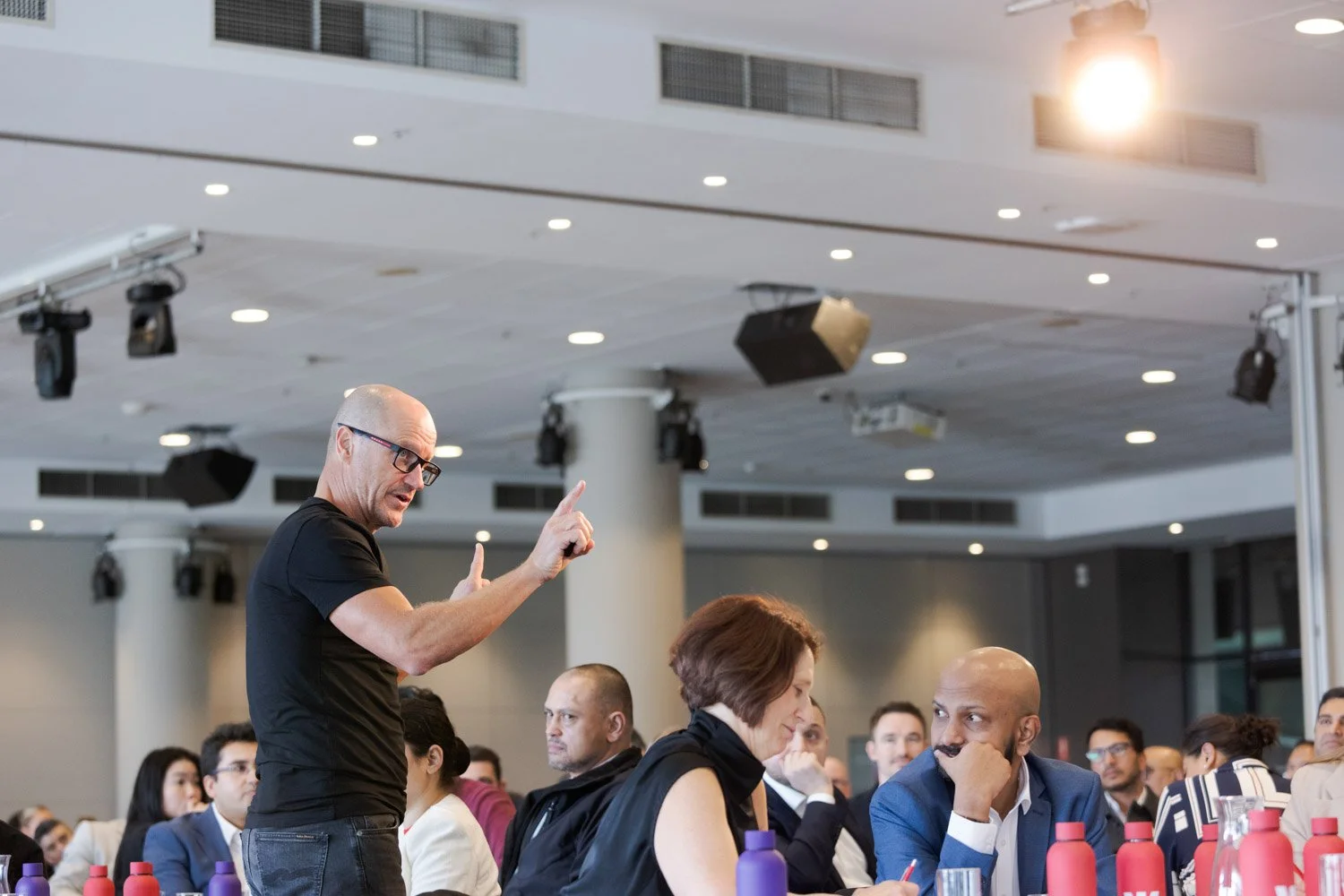 A man wearing glasses and a black shirt is speaking to a seated audience in a large conference room, gesturing with his. finger. The audience includes a woman with short hair and a man with a bald head, sitting at a table with purple and red water bo