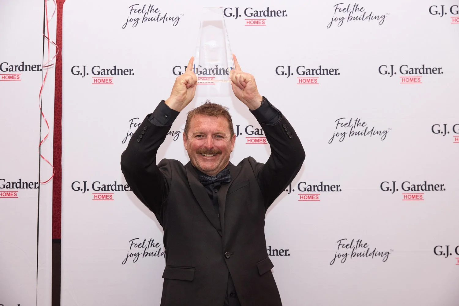 A man in a black suit and bow tie holding a clear award above his head, smiling at the camera, in front of a G.J. Gardner Homes backdrop.