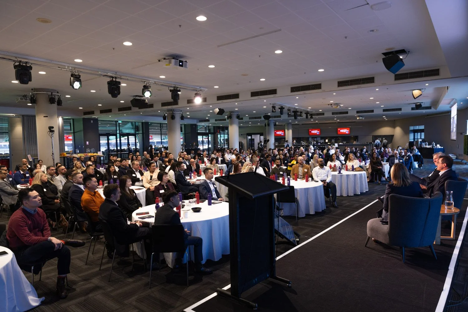 A large conference room filled with round tables and seated attendees listening to a speaker at a podium. The room has a modern design with ceiling lights, large windows, and multiple screen displays.