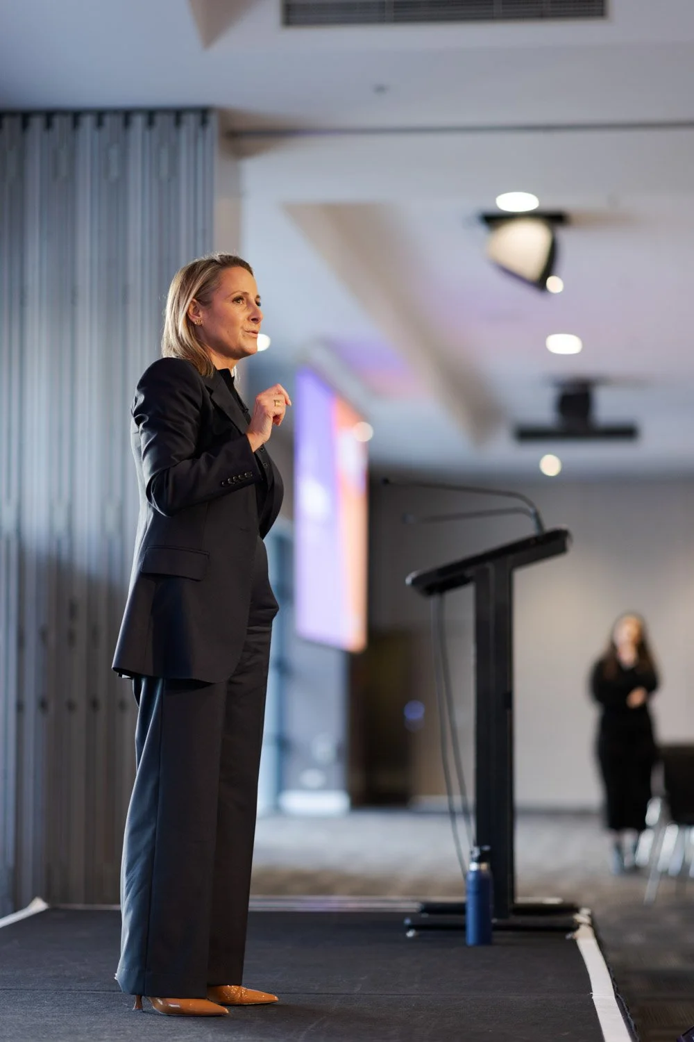 A woman in a black suit speaking at a conference podium.