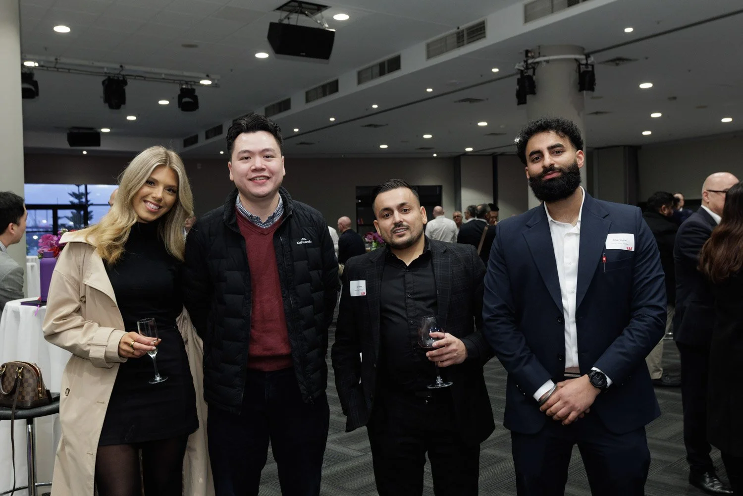Group of four diverse professionals at a formal event, standing together and smiling, with wine glasses in hand, in a modern conference room with other attendees in the background.