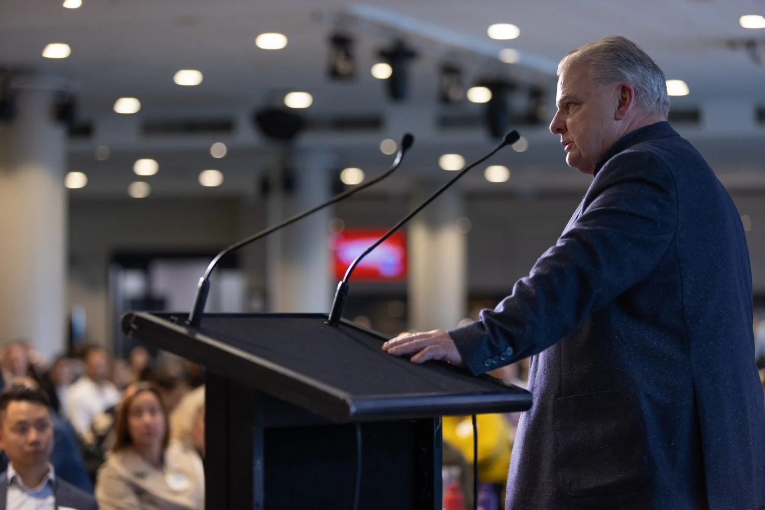 Man giving a speech at a podium in front of an audience in a conference room.