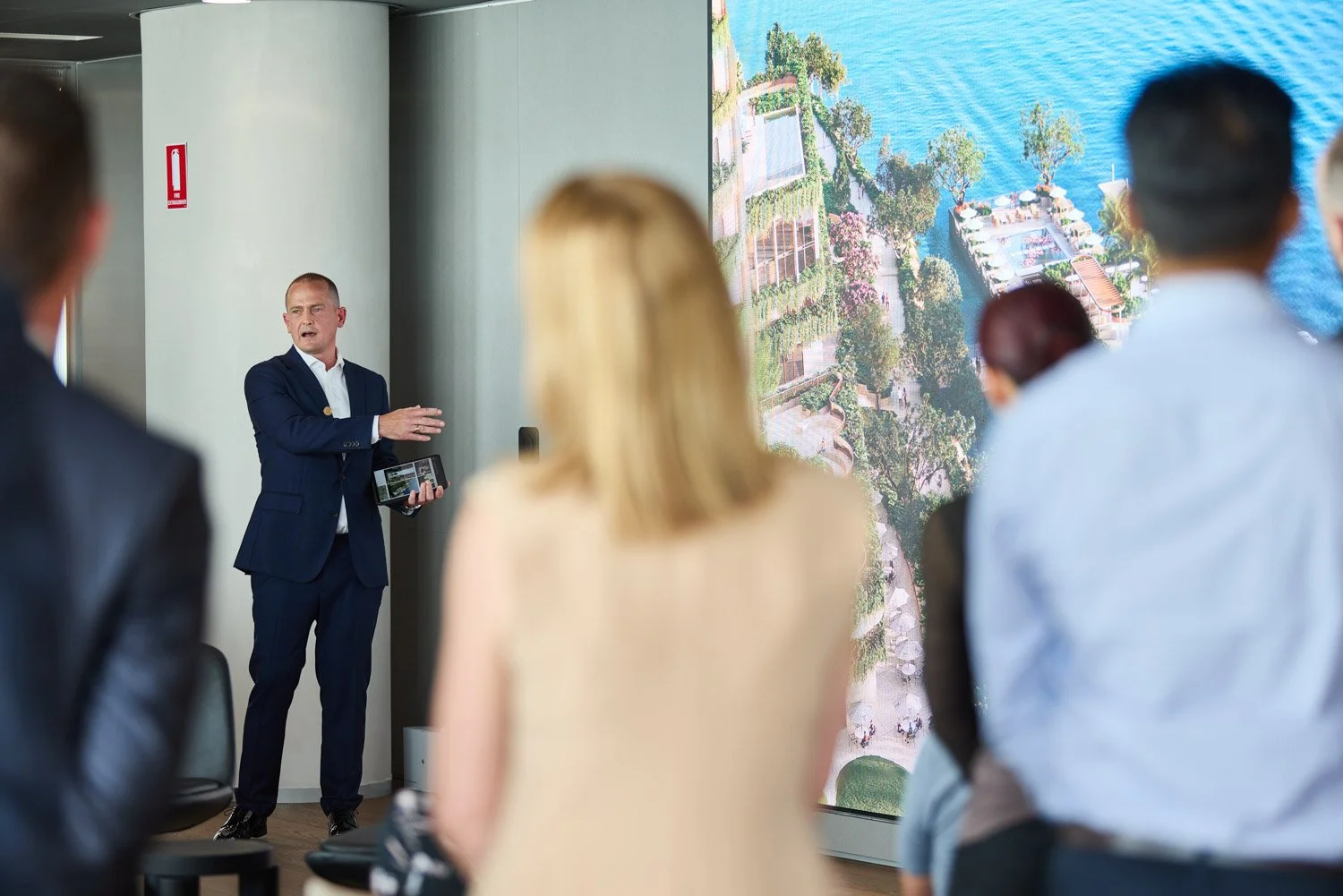 Man in a blue suit giving a presentation to an audience in a conference room, with a large screen showing an aerial view of a waterfront property.