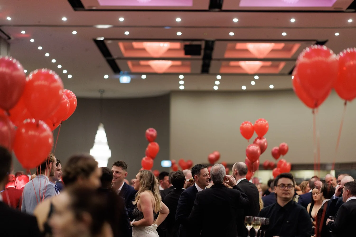 A crowded indoor event with people dressed in formal attire, red heart-shaped balloons floating above the crowd, and elegant lighting fixtures hanging from the ceiling.