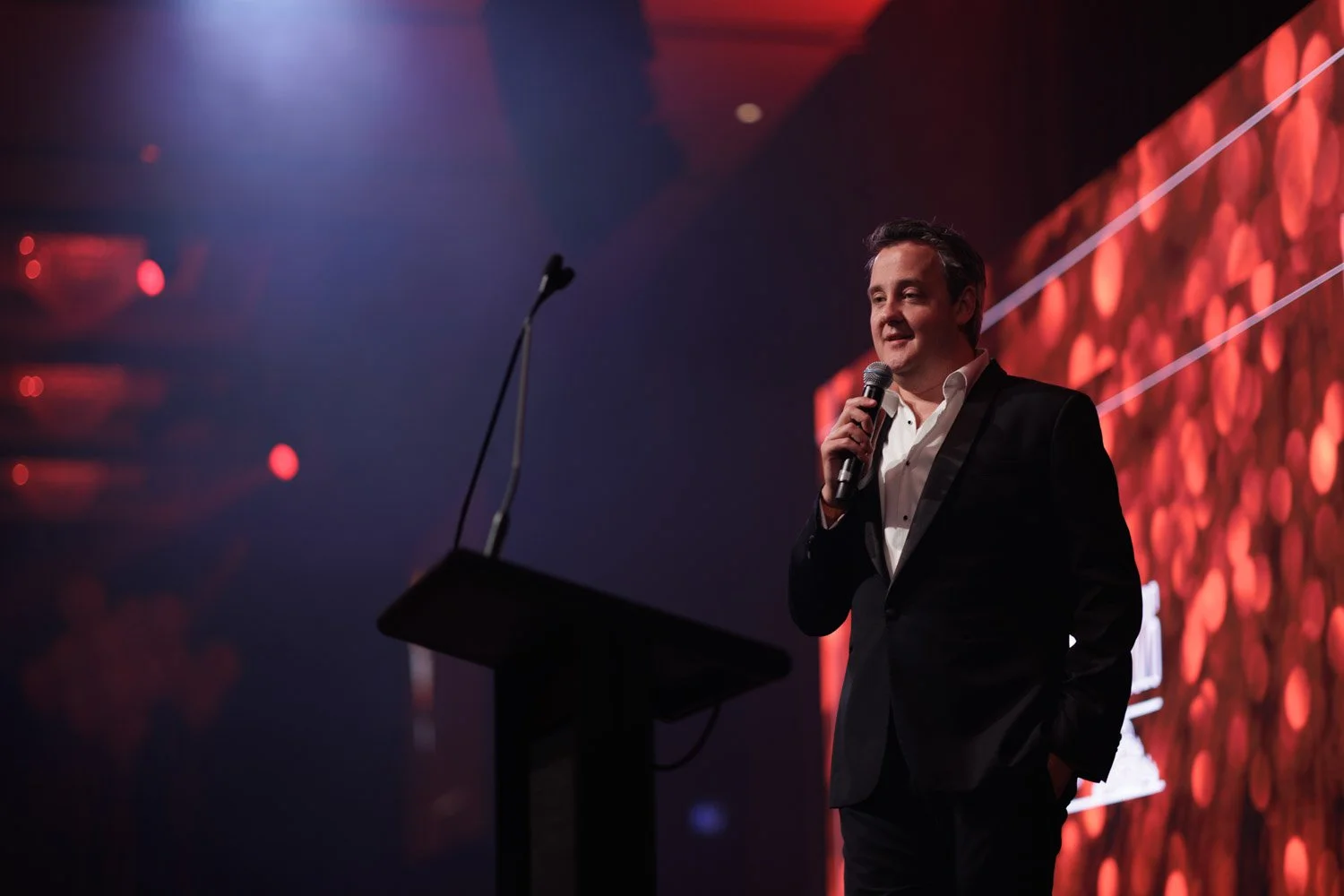 A man in a tuxedo speaking into a microphone on stage at a formal event with an illuminated background.