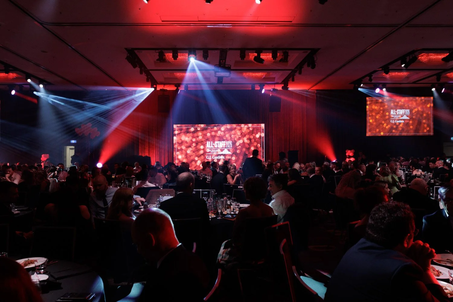 A large event hall filled with people sitting at round tables, with a stage at the front. The stage has a big screen displaying 'ALL-STAFF 2026' and the name G.J. Gardein, with dramatic red and white lighting and spotlights creating a festive atmosph