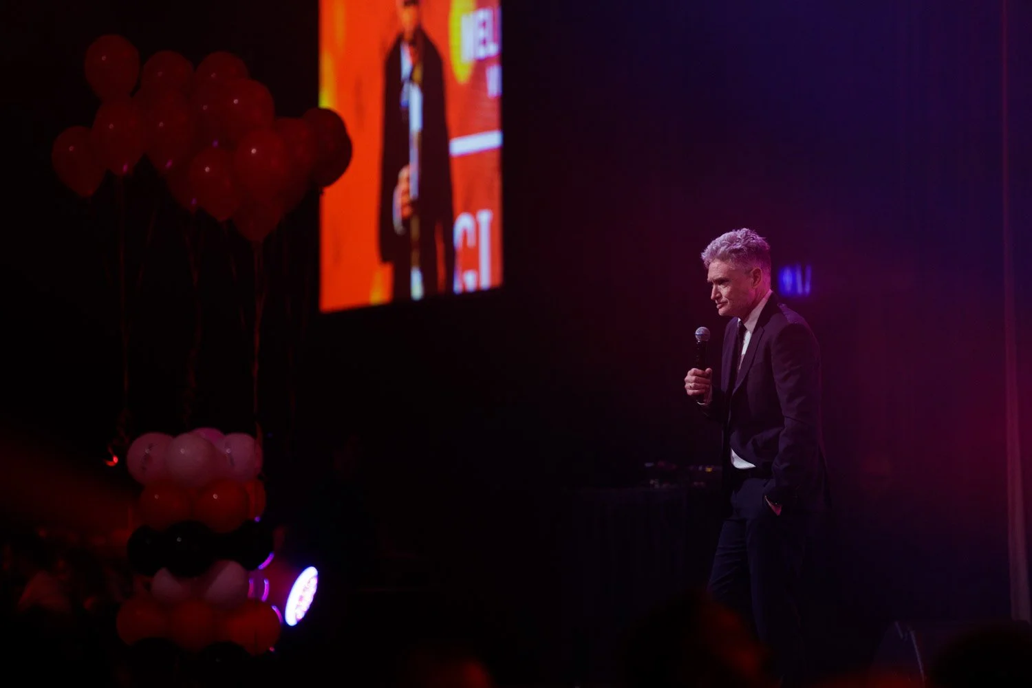 A man in a suit holding a microphone on stage during an event, with balloons arranged in vertical clusters on the left and a large screen displaying his image in the background.