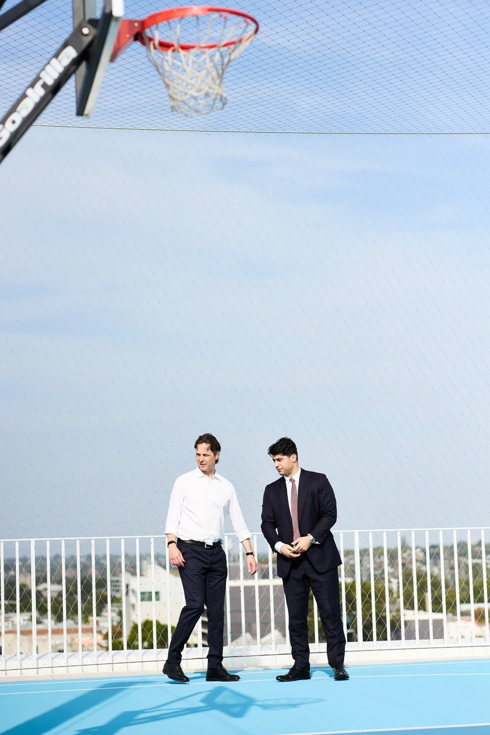 Two men in business attire walking on a rooftop basketball court beneath a basketball hoop with a net and blue sky