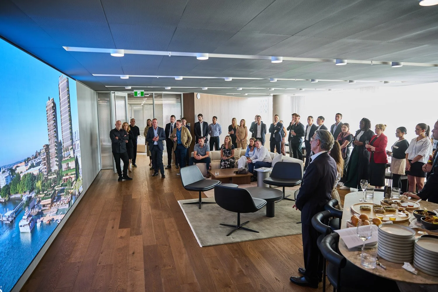 Business gathering in a modern conference room with a large screen displaying buildings and harbor, group of professionals standing and sitting, some with drinks, a man speaking at the front near a table with snacks and glasses.