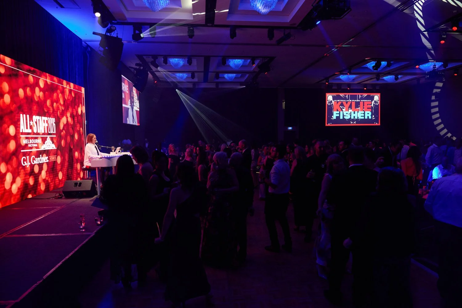 Crowd attending a formal event or conference in a large dark ballroom with stage, large screens, and colorful lighting, featuring a woman playing a piano on stage.