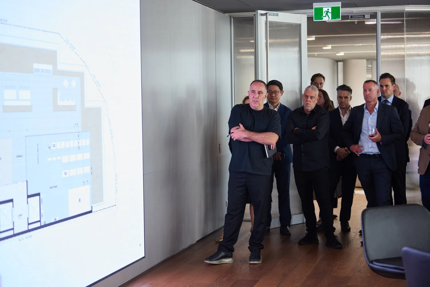 A group of people, mostly men in suits, standing in a modern conference room, listening to a speaker who is standing in front of a projection screen displaying a floor plan or blueprint. One man is holding a glass of wine. An emergency exit sign is v