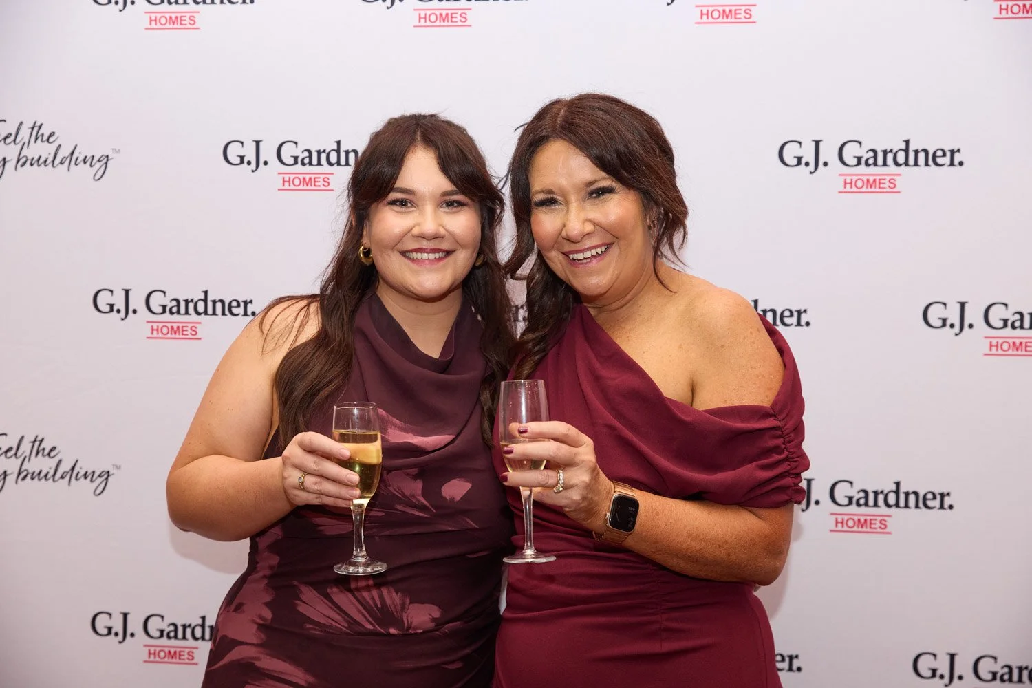 Two women smiling, holding glasses of champagne at an event with a G.J. Gardner Homes backdrop.