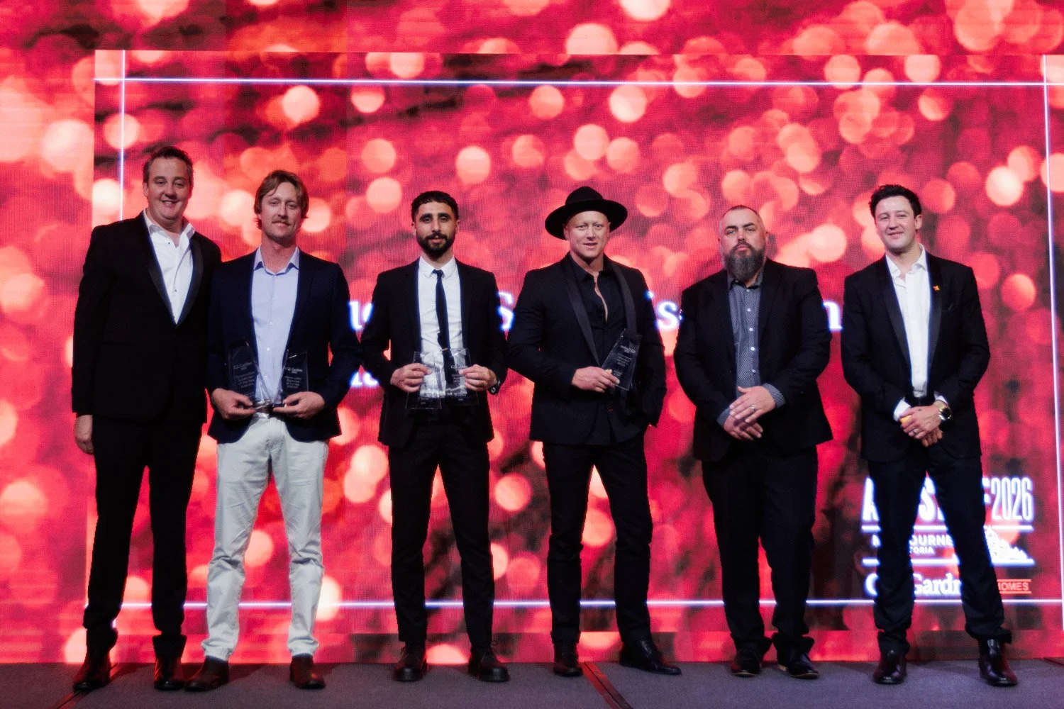 Six men in formal attire standing on stage at awards ceremony, holding glass awards, with a bright red and orange bokeh background.