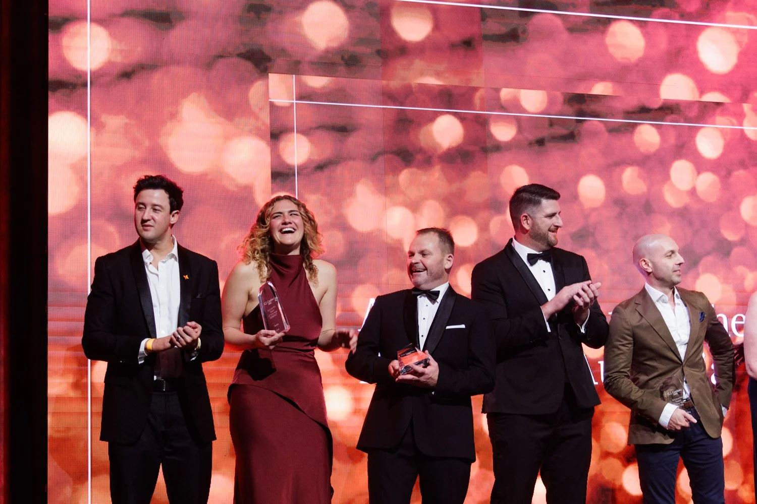 Six people in formal attire standing on stage, two holding awards, with a large, illuminated red and pink bokeh background, during an awards ceremony.