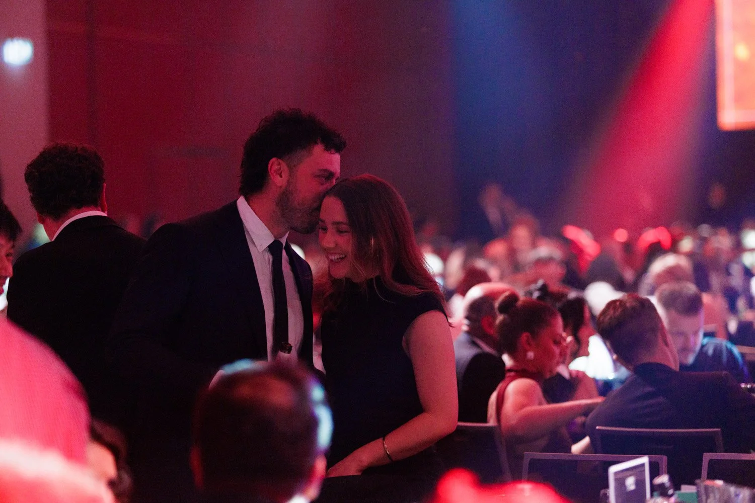 A man and woman dressed in formal attire sharing a tender moment, with the man kissing the woman's forehead, during a lively social event with many people in the background.